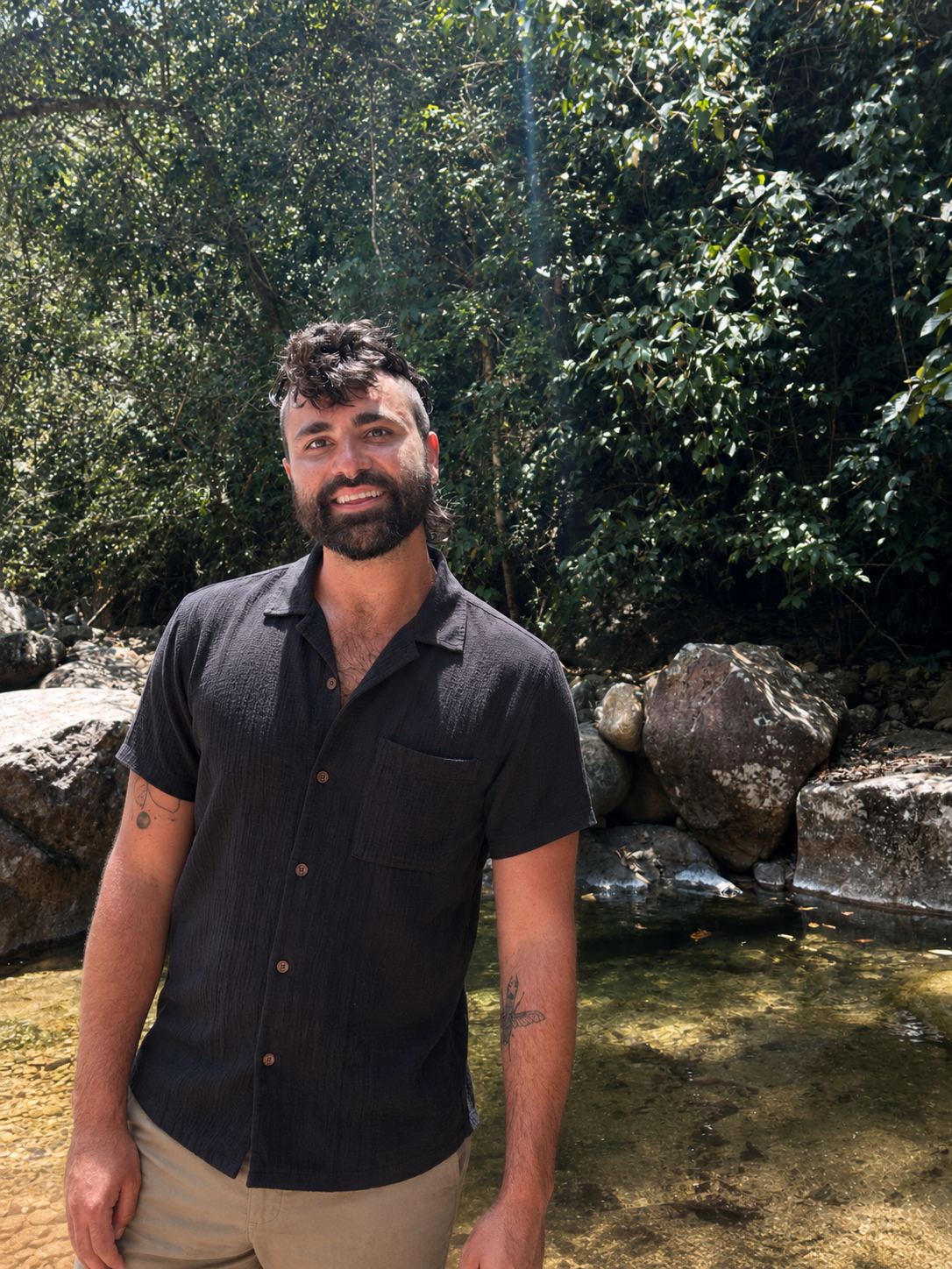 Spencer Smith standing in a shallow forest creek, smiling, with sunlit trees and mossy boulders behind him.